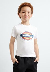 Balta medvilnė marškinėliai su apvaliu daugiaspalviu "Dickies" logotipu. Su juodomis sportinėmis kelnėmis, rankos kišenėse. Paprastas, kasdieniškas dizainas.