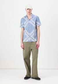 Camicia azzurra a maniche corte con motivo floreale bianco e pois, abbinata a pantaloni a gamba larga verde oliva e scarpe nere.