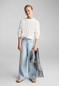 Weißer Kabelstrickpullover, hellblaue weitgeschnittene Jeans, beige Slipper und ein grauer Strickschal, der in der Hand gehalten wird. Glatte Texturen.