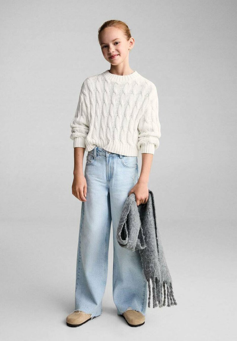 Weißer Kabelstrickpullover, hellblaue weitgeschnittene Jeans, beige Slipper und ein grauer Strickschal, der in der Hand gehalten wird. Glatte Texturen.