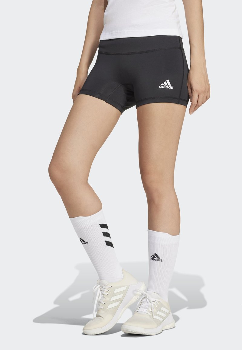 Pantaloni sportivi neri con una vestibilità aderente, caratterizzati da cuciture riflettenti e dal logo Adidas. Indossati con calze bianche e sneakers chiare.