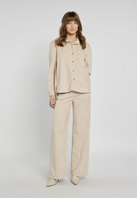 Beżowa koszula z korduroy z guzikami i kieszonką na piersi, w połączeniu z szerokimi spodniami w tym samym kolorze. Gładka tekstura, minimalistyczny design i dopasowany krój.
