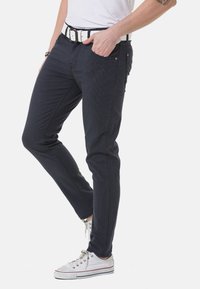 Dunkelblaue Jeans mit kariertem Muster, schlanker Schnitt, Seitentaschen und einem weißen Gürtel. Getragen mit weißen Turnschuhen.