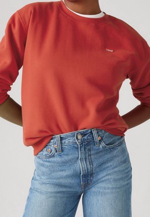 Person iført en knaldrød langærmet sweatshirt med Levi’s-mærke og lyseblå højtaljede jeans, hænderne hvilende på hoften mod en neutral baggrund.