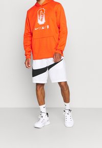 Sweat à capuche Nike orange avec logo gris, associé à un short blanc présentant une bande diagonale noire. Une paire de chaussures de sport noires et blanches complète la tenue.