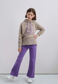 Beige Kapuzenpullover mit lila Totenkopf-Graphic, kombiniert mit lila Schlagjeans. Modell trägt weiße Sneakers, vor einem neutralen Hintergrund.