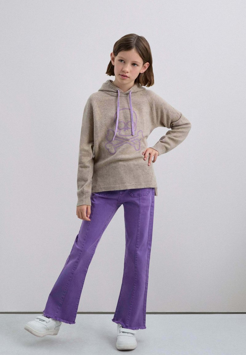 Beige Kapuzenpullover mit lila Totenkopf-Graphic, kombiniert mit lila Schlagjeans. Modell trägt weiße Sneakers, vor einem neutralen Hintergrund.