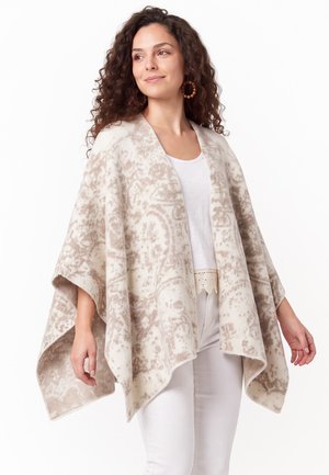 Junge Frau mit lockigem Haar, die große Creolen trägt und einen beigefarbenen gemusterten Poncho über einem weißen Oberteil und weißen Hosen trägt, die zur Seite schaut.