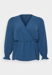 Blouse enveloppante bleue en tissu texturé. Dotée de longues manches bouffantes avec des poignets élastiques, d'une bordure en dentelle au niveau du décolleté et d'une taille cintrée.