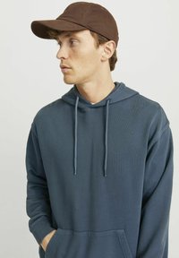 Sudadera azul con cordones y un bolsillo delantero, combinada con una gorra marrón. La sudadera presenta una tela texturizada y un corte holgado.