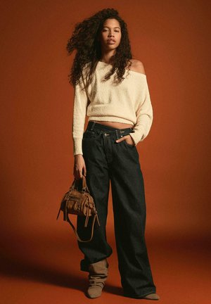 Femeie cu păr lung și creț, purtând un pulover crem cu umerii goi, blugi largi închiși la culoare, cizme maro deschis, ținând o geantă maro pe fundal maro.