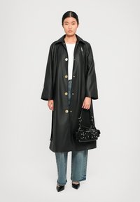 Cappotto trench in pelle nera con bottoni dorati, vestibilità comoda, fessure laterali, indossato sopra una top bianco, abbinato a jeans blu chiaro e una piccola borsa nera.