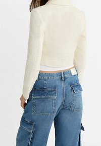 Femme portant une veste blanche courte texturée et un pantalon cargo en denim bleu taille haute avec de grandes poches à rabat, vue de dos.