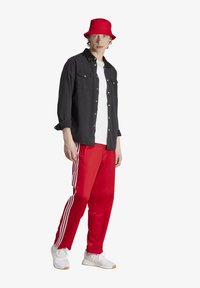 Schwarze Button-Up-Hemd, weißes T-Shirt, rote Jogginghose mit weißen Streifen, roter Bucket Hat und weiße Sport Schuhe mit beigen Akzenten.