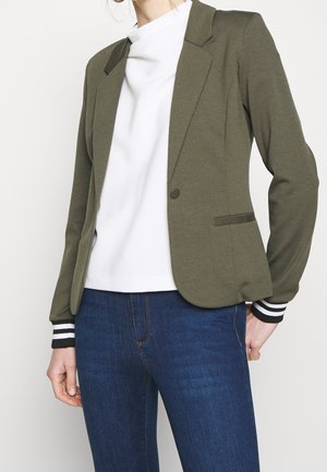 Woman wearing olive green blazer with single button, white top, and blue jeans, standing with one hand on hip against plain background.