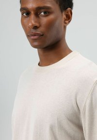 Beige trui gemaakt van zachte stof met een ronde hals en geribbelde randen. Gladde textuur, getoond op het bovenlichaam van een model, neutrale achtergrond.