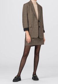 Blazer marron à carreaux avec accents noirs aux poignets, assorti à une jupe coordonnée, collants noirs transparents et mocassins noirs ornés d'une chaîne dorée.