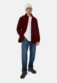 Ung mand iført en hvid beanie, hvid t-shirt, bordeauxfarvet fløjlester, blå jeans og sorte sneakers, der står foran en ensfarvet baggrund.