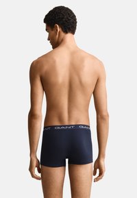 Mann von hinten gesehen, trägt dunkelblaue GANT Boxershorts, steht vor einem schlichten hellen Hintergrund.
