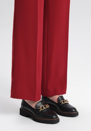 Black leather loafers with a gold chain accent, featuring a sturdy sole and smooth texture, paired with wide red trousers.