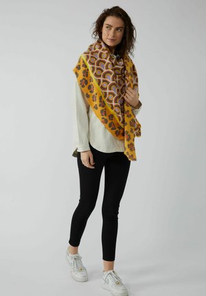 Écharpe jaune avec imprimé léopard brun et motifs circulaires colorés, drapée sur une chemise claire, associée à un pantalon noir et des baskets blanches.