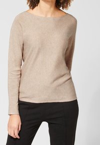 Femme portant un pull beige à manches longues avec un décolleté bateau et un pantalon noir ajusté, se tenant devant un fond clair uni.