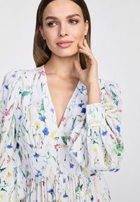Sukienka w kwiatowy print z głębokim dekoltem w kształcie litery V, bufiastymi rękawami i warstwowym dołem. Kolory to biały, zielony, niebieski, różowy i żółty.
