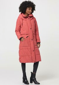 Lange roze pufferjas met een capuchon, voorzien van een ceintuur in de taille, drukknoopsluiting, gewatteerde textuur en voorkeurszakken. G gedragen over zwarte jeans en laarzen.