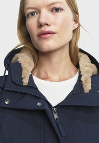 Mlada žena svijetle puti i plave kose nosi tamnoplavi kaput s bež umjetnim krznom i bijelu majicu ispod.