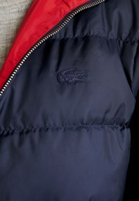 Chaqueta acolchada azul marino con cuello de acento rojo, cierre con cremallera y una tela texturizada. Presenta un logotipo bordado en la parte izquierda del pecho.