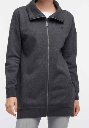 Donna che indossa una giacca in pile con cerniera di colore grigio scuro, con collo alto e tasche laterali, abbinata a pantaloni grigio chiaro.