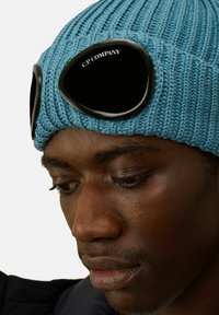 Cappello di lana blu lavorato a maglia con una texture a coste e occhiali neri attaccati, con il logo "C.P. COMPANY" sul bordo frontale.