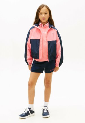 Ragazza in piedi che indossa una giacca rosa e blu navy, una felpa rosa, pantaloncini blu navy, calzini bianchi e sneakers blu navy su sfondo bianco.