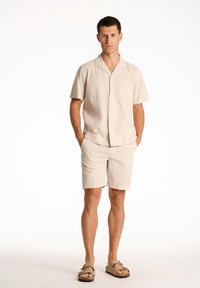 Vaalean beige lyhythihainen napillinen paita, jossa on tekstuuri, yhdistettynä samanvärisiin shortseihin. Mallilla on jalassaan litteät sandaalit, kädet taskuissa.