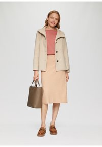 Beige wollen jas met een hoge kraag en bruine knopen, gecombineerd met een roze top, beige kokerrok, bruine tas en leren schoenen.