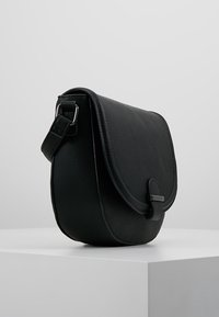 Borsa a tracolla nera in pelle con chiusura a pattina e tracolla regolabile, posizionata su un piedistallo bianco contro uno sfondo grigio uniforme.