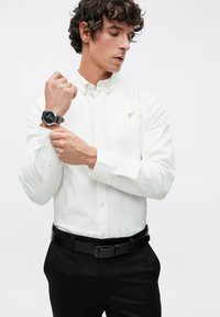 Weißes Hemd mit Knopfleiste und dezentem Logo, schwarze Anzughose und eine schwarze Uhr. Glatter Stoff, figurbetontes Design.