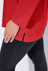 Rode longsleeve top met een afgeronde zoom en zijsplitten, gemaakt van zachte stof. Draag het met donkere spijkerbroeken, met een close-up van de mouw.