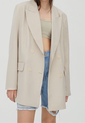 Frau trägt beigefarbenen zweireihigen Blazer über beigem Croptop und ausgefransten Jeansshorts vor schlichtem weißen Hintergrund.