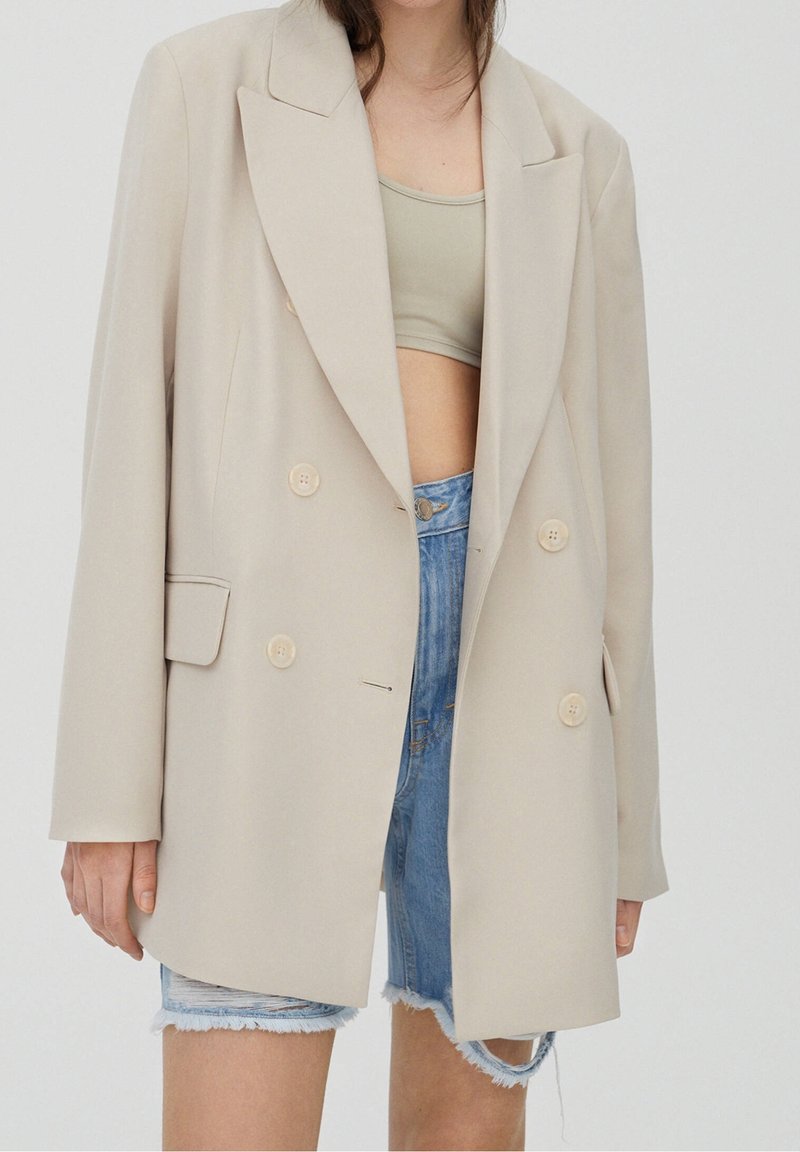 Femme portant un blazer croisé beige sur un crop top beige et un short en jean effiloché sur fond blanc uni.