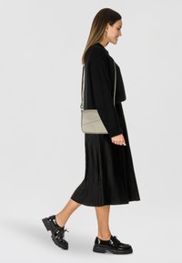 Bolso cruzado en gris claro, con un diseño estructurado, textura suave y ligeros acentos geométricos. Combinado con una falda negra plisada y zapatos.