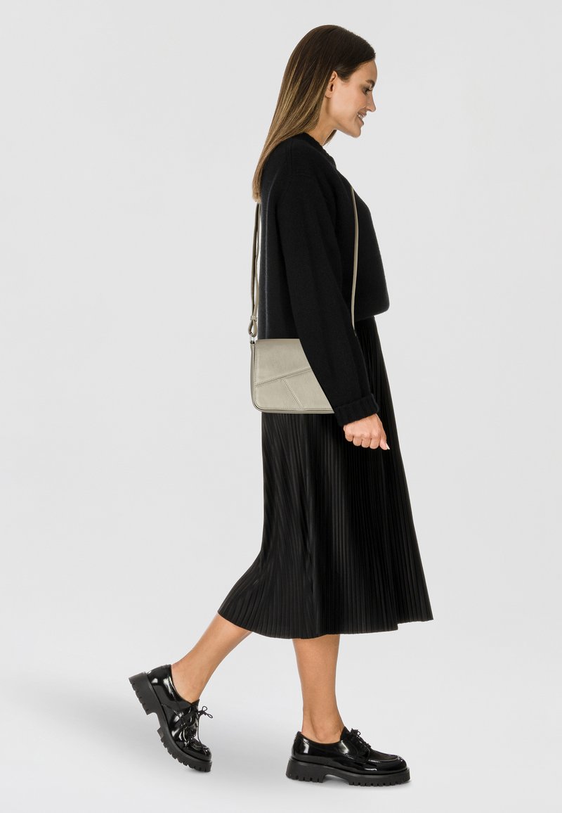 Bolso cruzado en gris claro, con un diseño estructurado, textura suave y ligeros acentos geométricos. Combinado con una falda negra plisada y zapatos.