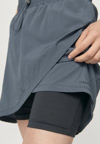 Lichte, grijze hardloopshorts met een elastische tailleband en trekkoord, gedragen over zwarte compressie-shorts. Glad materiaal, minimaal merkdesign.