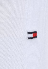 Baltie kokvilnas zeķes ar nelielu izšūtu logo, kas sastāv no sarkanām, baltām un melnām taisnstūrām. Audumam ir gluda tekstūra ar ribotu detaļu.