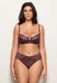 Donna che indossa un reggiseno e delle mutandine in pizzo viola scuro abbinati, in piedi contro uno sfondo chiaro e semplice, rivolta in avanti con espressione neutra.