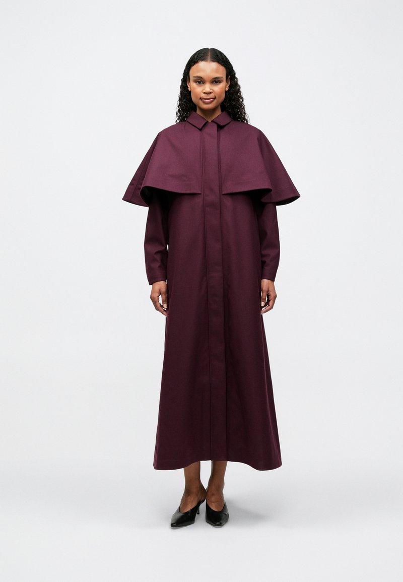 Sieviete ar cirtainiem matiem stāv, ģērbta garā, tumši violetā mētelī ar apmetni un melnām, punta pirksta kurpēm uz viendabīga fona.