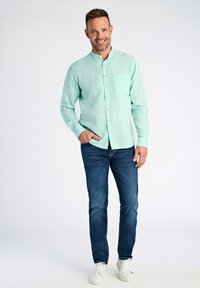 Camicia button-up di un leggero blu teal con taschino sul petto, abbinata a jeans blu scuro e sneakers bianche. Tessuto a trama, vestibilità rilassata.