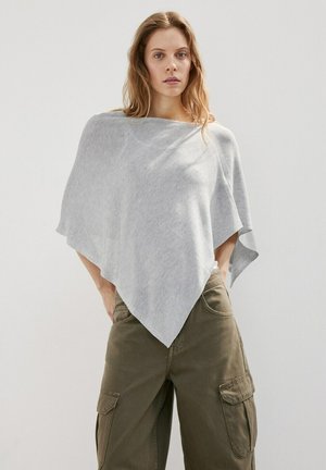 Žena v svetlosivej asymetrickej pončo a olivovo zelených cargo nohaviciach, stojaca s rukami v vreckách na jednofarebnom pozadí.