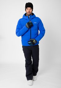 Blue puffer jacket with a hood, zippers, and insulation. Worn with navy pants and gloves featuring gray and green accents. White sneakers.