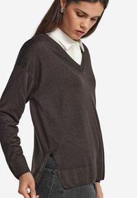 Brauner V-Ausschnitt-Pullover mit langen Ärmeln, seitlichen Schlitzen und gerippten Bündchen. Leichter Strickstoff, der über einem weißen Hemd mit Kragen getragen wird.